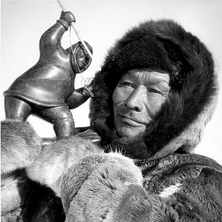 Black and white close-up photo of a man in a parka and fur mittens holding a small sculpture of a man with a harpoon.