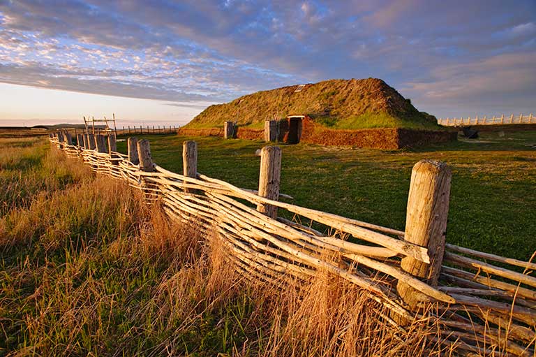 This image shows a wooden fence close to the camera and in the distance a Norse building on a blue and cloudy sky. 