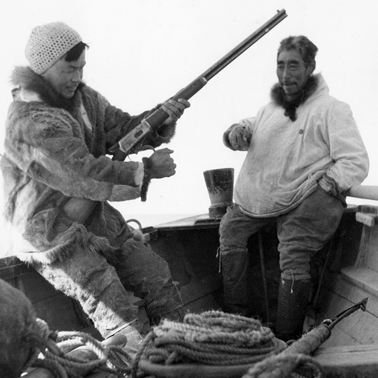 Black and white photo of two men in parkas sitting on the edge of the inside of a boat. The man on the left holds and inspects a rifle.
