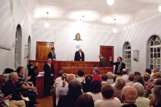 An audience sits in a courtroom, while six people dressed in historic costumes act out a trial.