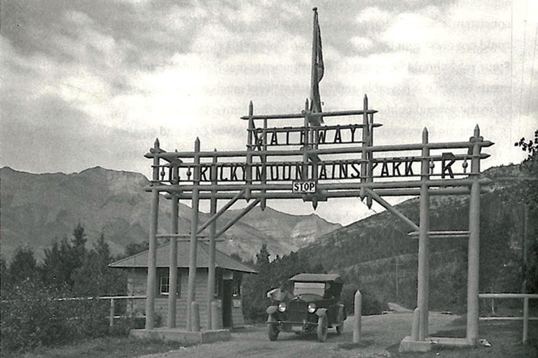 Mountains in the background, in the foreground a large wooden gate, a booth, and a man leaning against his old car posing for the photo. 