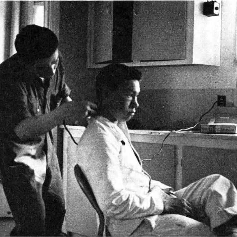 Black and white photo of one man sitting in a chair while a man behind him trims his hair with an electric razor.