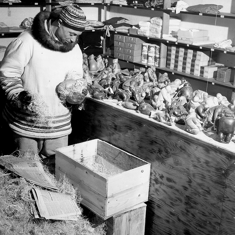 Black and white photo of a man in a parka and hat holding a small sculpture wrapped in protective fibres. To the right of the man is a bench with numerous other small sculptures awaiting packaging.