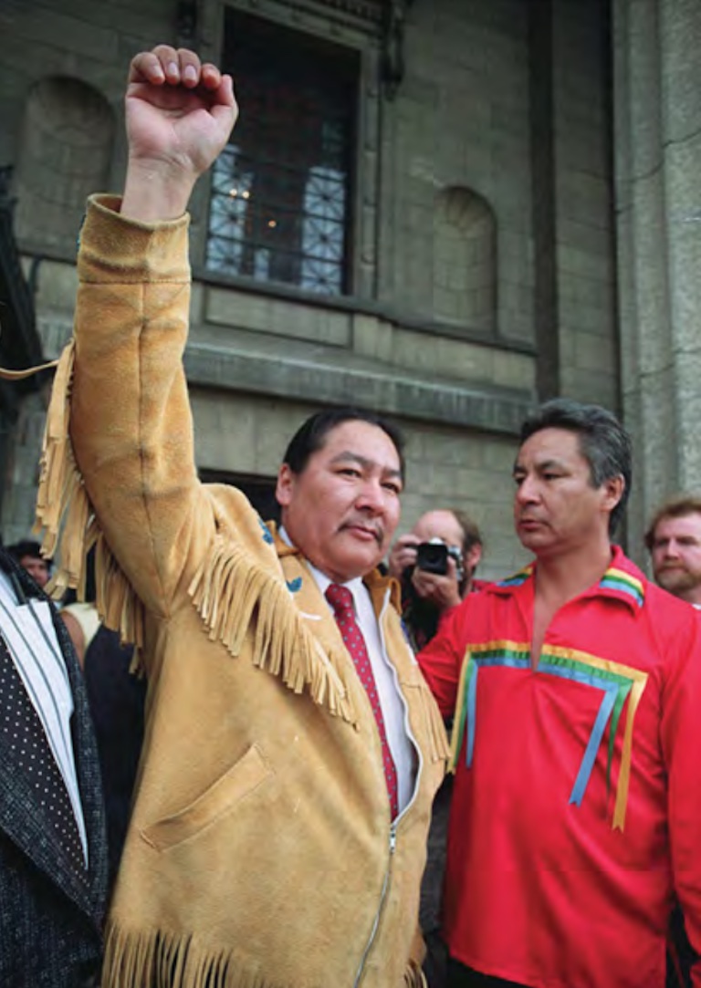 A man in a leather fringe coat stands with his hand outstretched in the air in a fist. A man in a red shirt stands beside him on the right.