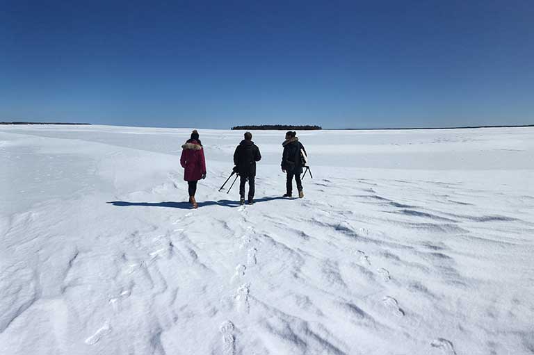 Snow landscape and bright blue sky with three people standing side by side facing away from the camera. 