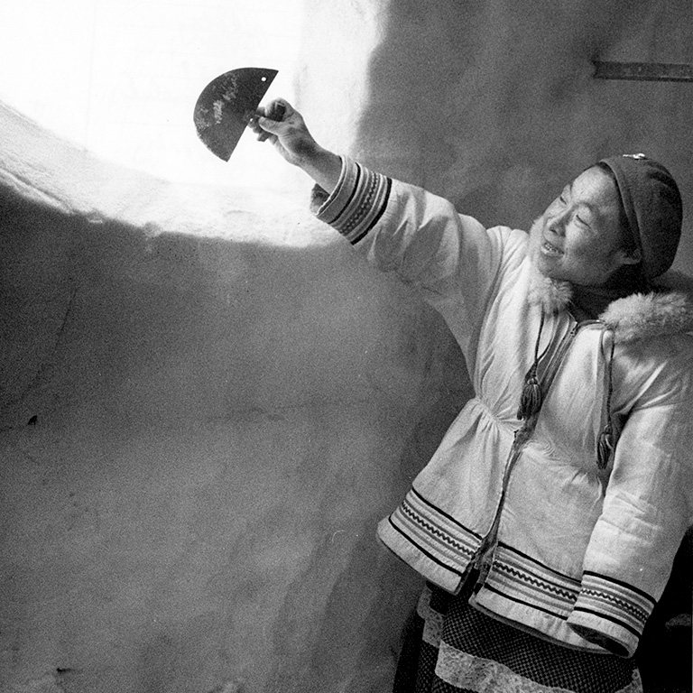 Black and white photo of a woman in a parka and hat holding a semi-circle shaped knife and pointing it at an open window cut into an igloo.