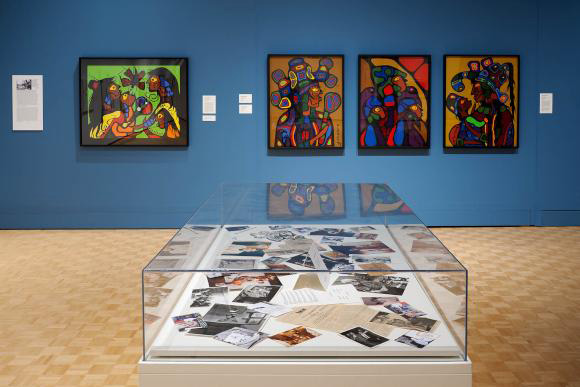 The interior of an art gallery showing a glass display case with objects and a blue wall with four paintings on it.
