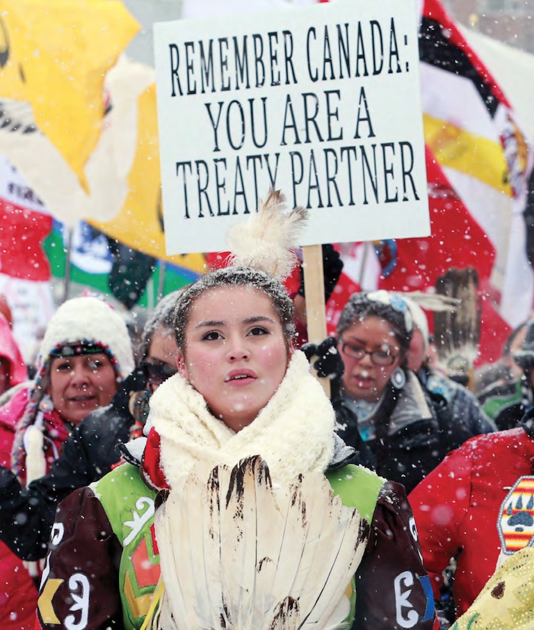 A woman stands with protesters holding signs. Everyone wears winter apparel and it is snowing. The young woman holds feathers and has a feather in her hair.