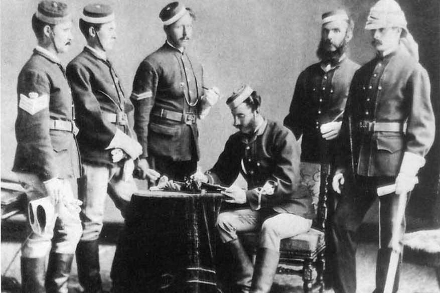 A man sits at a table inspecting a letter while five other men stand around him watching, two on the right and three on the left. 