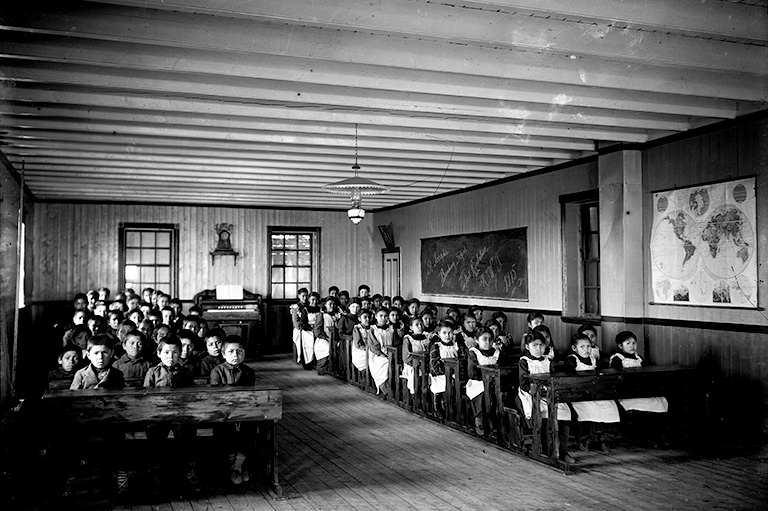 Black and white photo of students in uniforms sitting behind desks in a classroom.