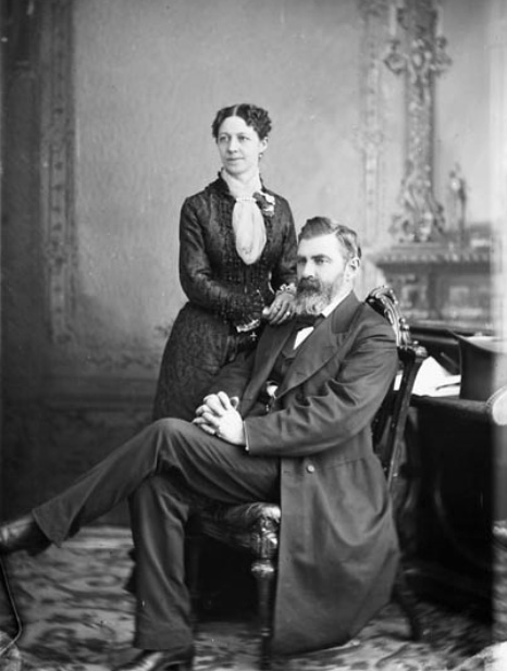 A photograph of a man sitting in a chair with his legs crossed and a woman standing on his right with her hands on his shoulder. 
