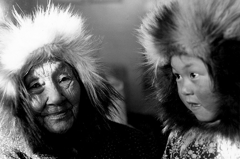 A grandmother with a fur parka framing her face sits beside a child who also wears a fur parka