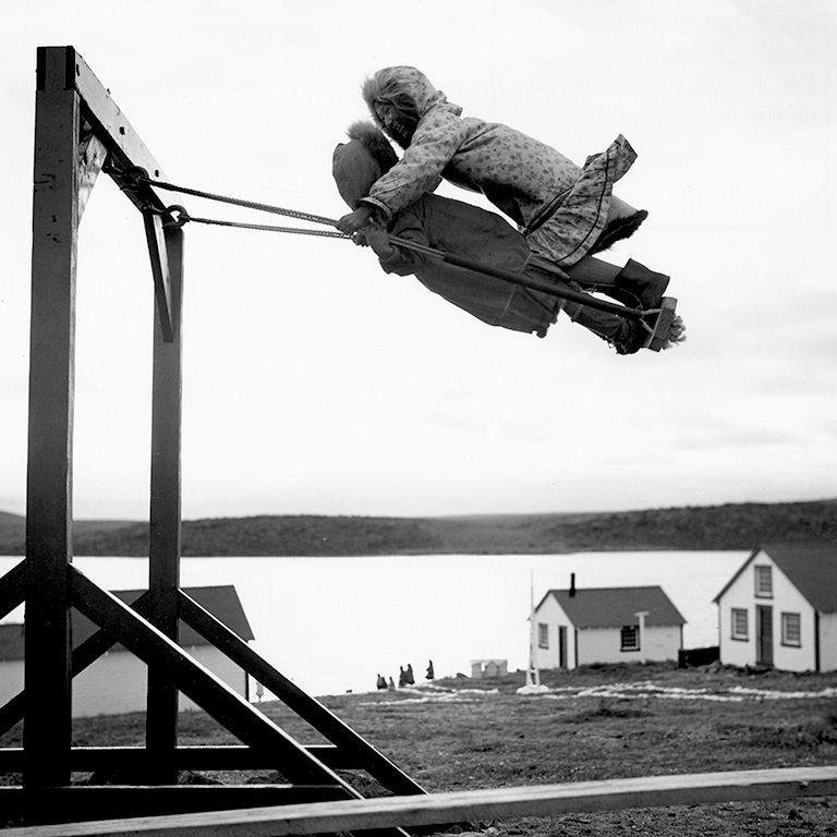 Two girls in parkas stand on a swing as it is midway through the air.