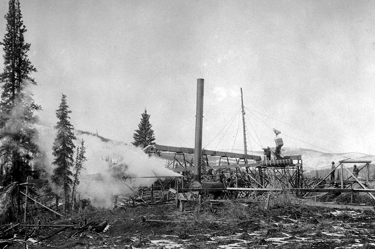 Black and white photo of sprawling machinery in front of trees with steam coming out of a pipe of one machine.