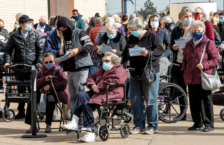 Seniors in Canada gathered wearing masks. Some people are standing filling out paperwork to get vaccinated. There is a woman seated in a wheelchair at the front of the group.