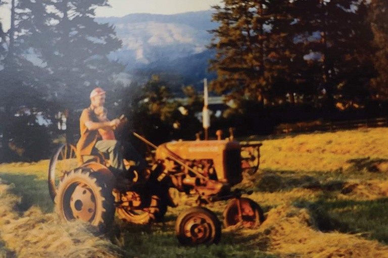 A young girl sits on her grandfather's lap riding a tractor.