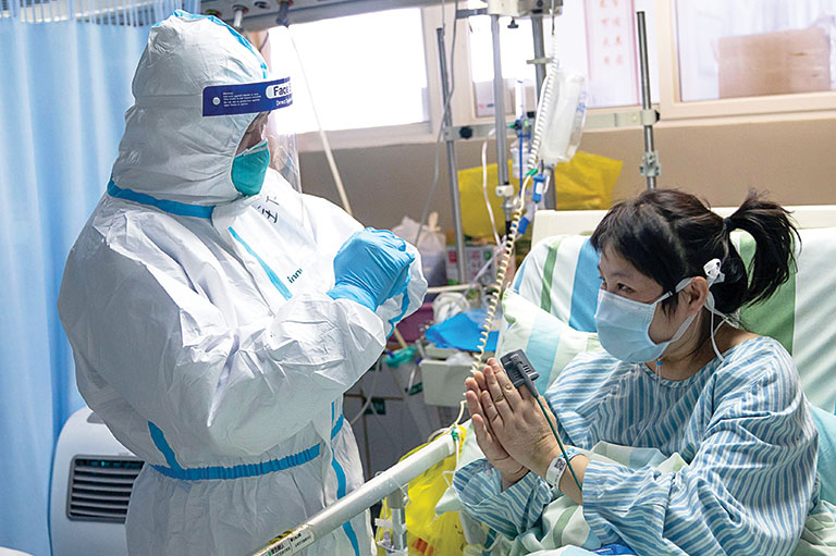 A person in full PPE treats a patient in a hospital bed wearing a mask. The patient holds their hands up to thank the medical worker.