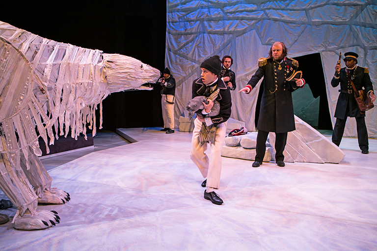 Image shows three people on stage walking fearfully around a large polar bear. The polar bear is made of art supplies. 