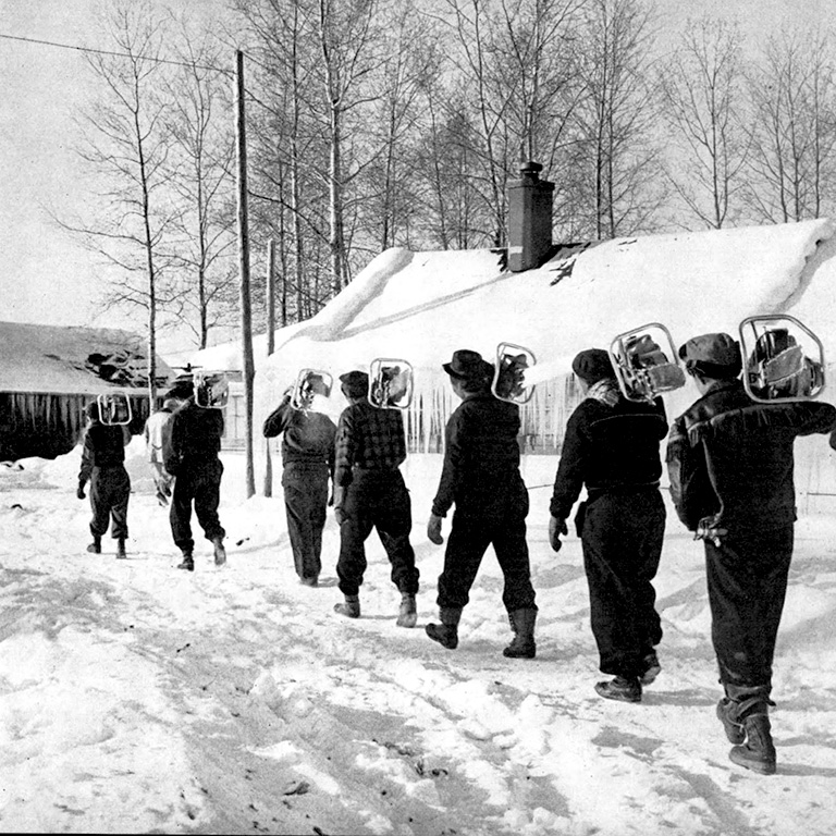 Black and white photo of seven men walking away from the camera in the snow carrying chainsaws over their shoulders.