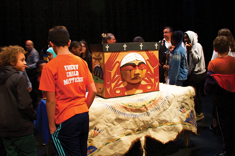 Students in orange shirts look at a wooden box with a mask on the front. The box is on top of a table covered in a table cloth.