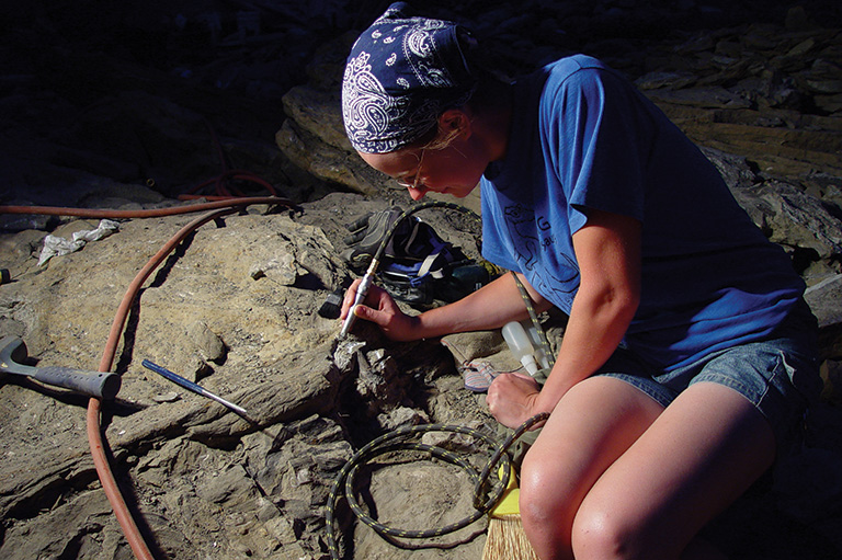 A lady in shorts and a blue tshirt sitting on the ground leaning over the work site. 