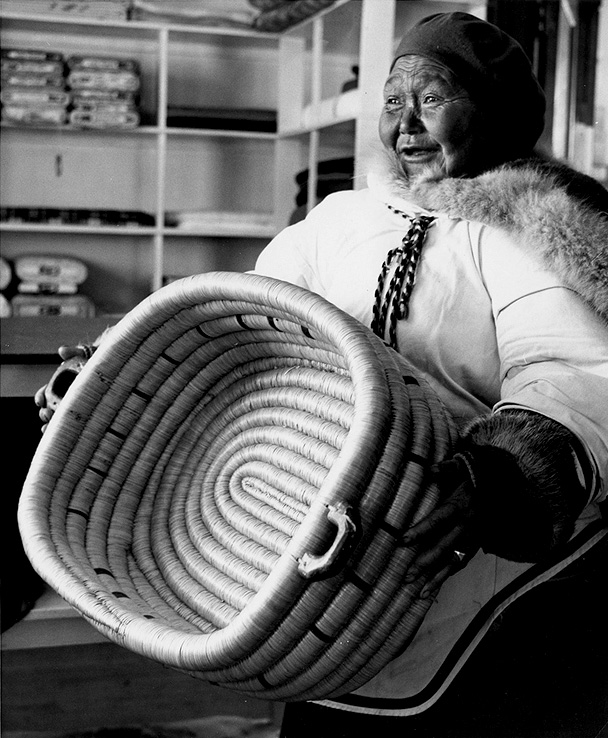 Black and white photo of a woman in a parka and hat holding a large woven basket in front of her.