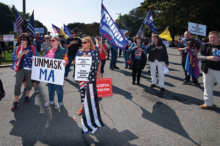 Protesters march with flags and signs that read "Unmask MA" and "Trump."