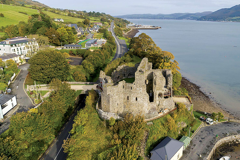 An aerial view of a large stone castles surrounded by grass and trees, but close to the water on the right side of the image.