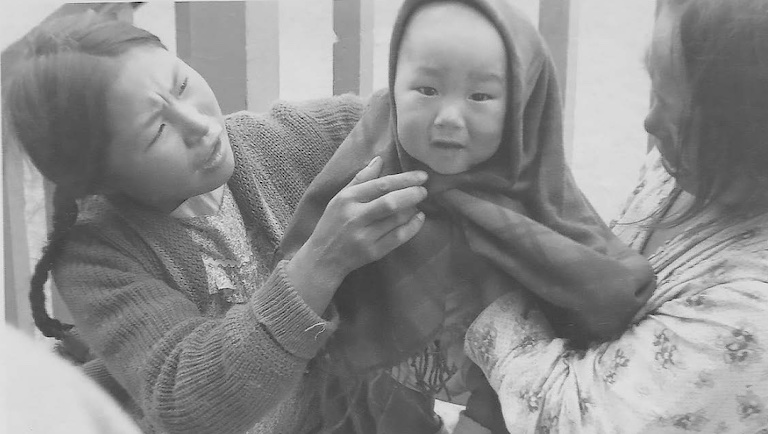 Black and white photo of two Inuit women holding a baby in between them. The baby smiles at the camera and has a blanket around it.