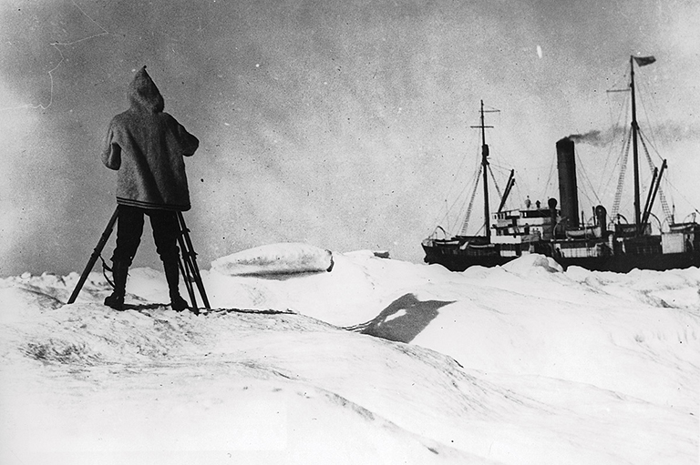 This is a greyscale image that shows a ship in the distance and a person in a coat in the foreground.