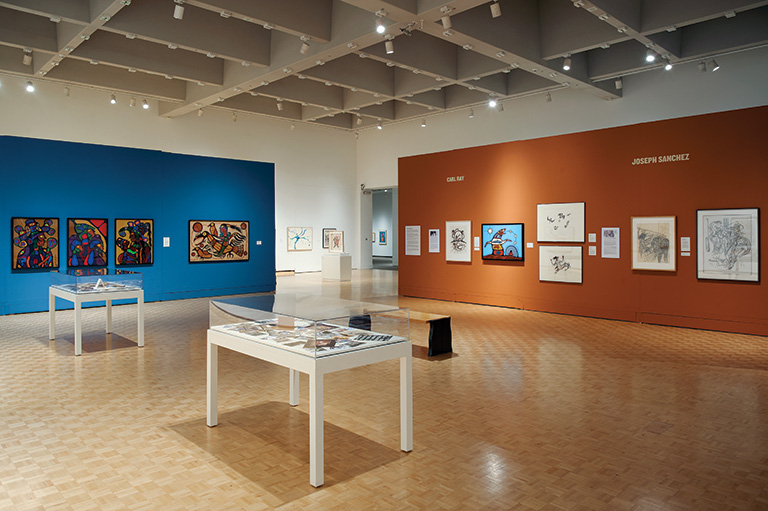 Interior shot of an art gallery with one blue wall and one brown wall. There is a white table with a glass top in the middle of the room.