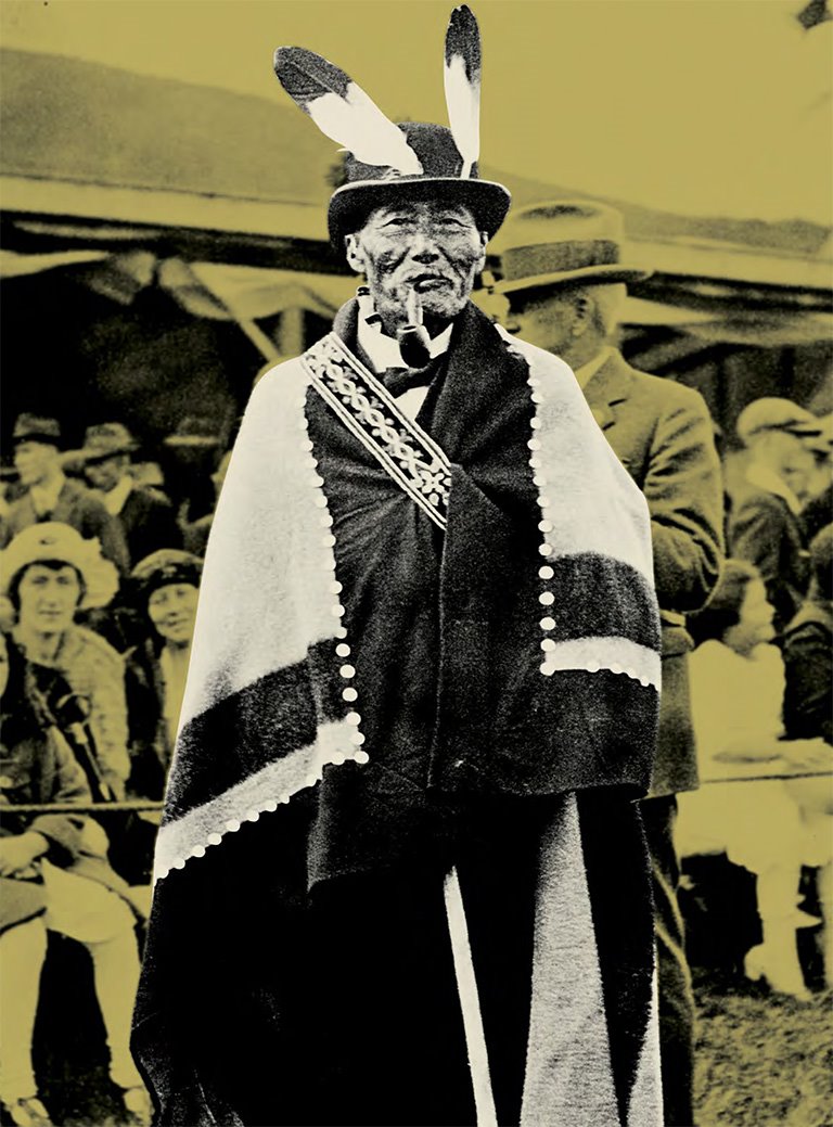 A man in a hat with two feathers on the front has a striped blanket around his shoulders. The photo is edited so that the background has a yellow tint.