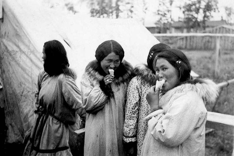 Black and white photo of four women in parkas. Two are eating ice cream and looking towards the camera.