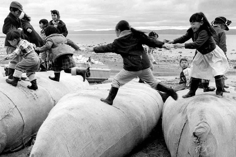 A black and white photo of children in coats and boots running across large supply sacks.