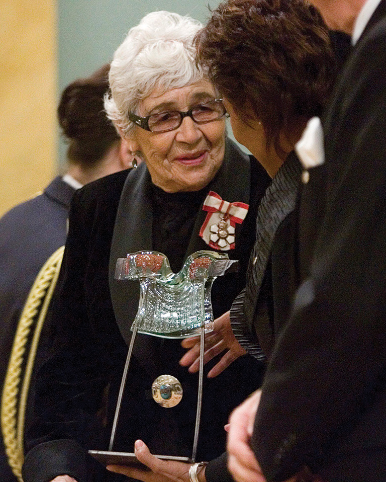 A woman with white hair and glasses speaks to a woman with brown hair holding a glass award.