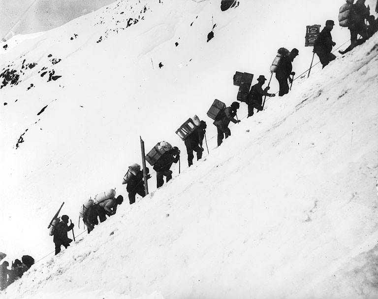 A black and white photo shows prospectors with large backpacks hiking a portion of a snowy mountain.