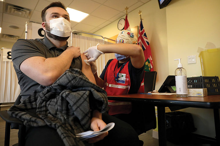 A man in a grey collared t-shirt sits in a chair in a mask while a person in medical scrubs and apparel gives him a vaccine.