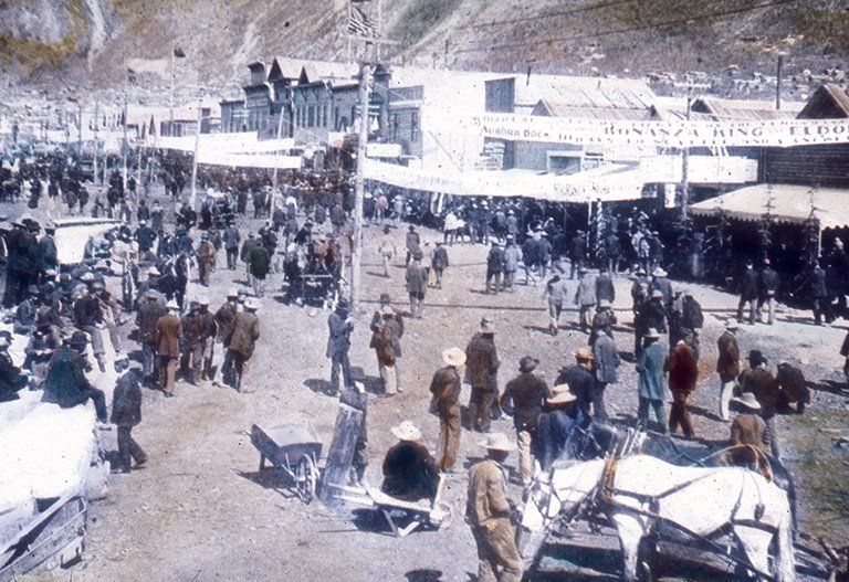 Photo of people walking the dirt streets of a city with a row of stores in the background and two horses in the foreground.