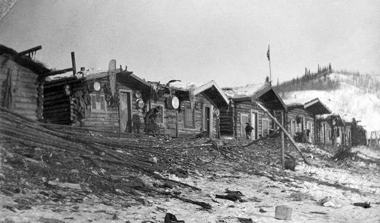 Black and white photo of a row of six log cabins in front of a snowy mountain.