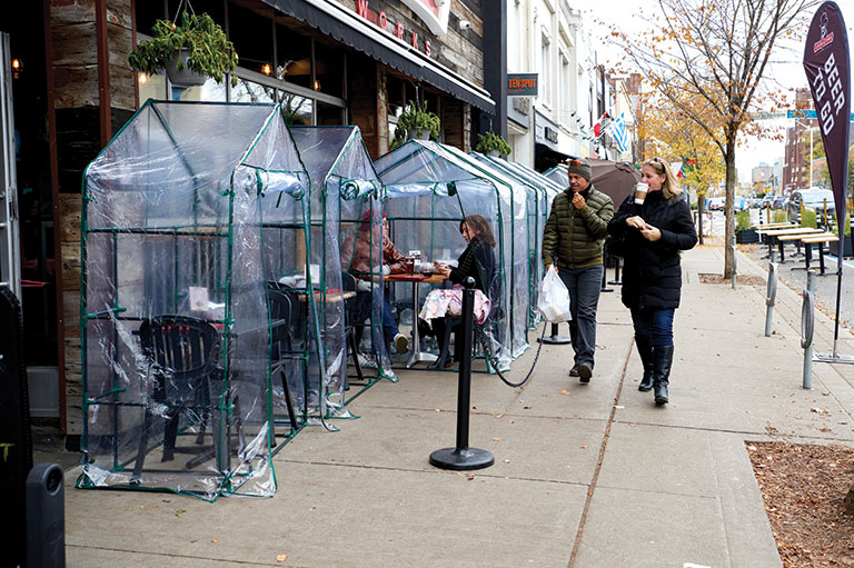 See-through plastic tents are setup around dining tables on a sidewalk patio.