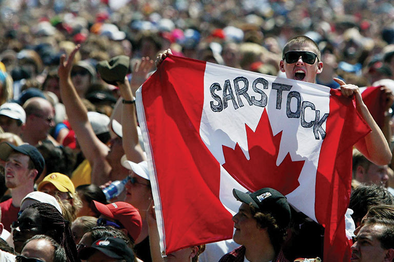 A large crowd is gathered for a concert. Two concert-goers hold a Canadian flag with the word "Sarstock" written across the top.