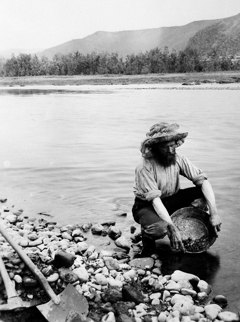 Black and white photo of a man in a hat kneeling in shallow water with a pan in front of him that has rocks and water in it.