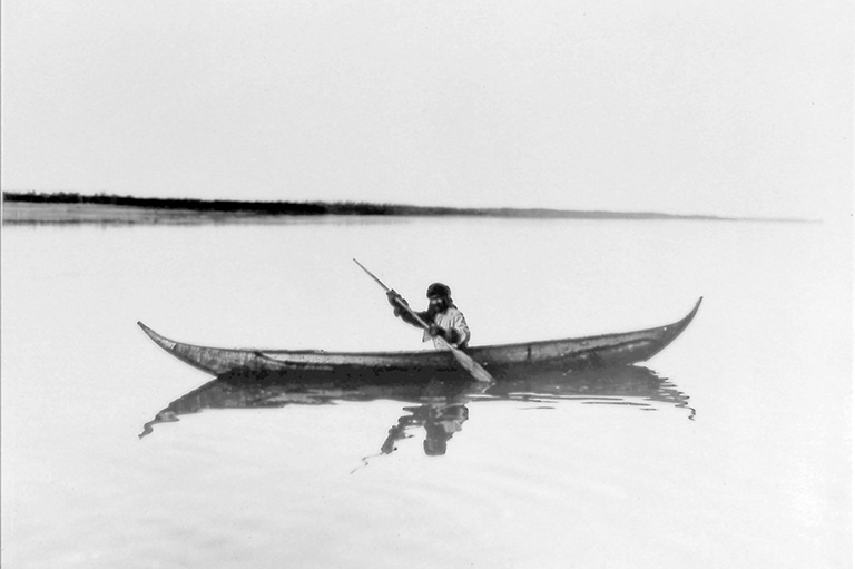 Black and white photo showing a lone man paddling in a canoe wearing a fur hat and gloves.