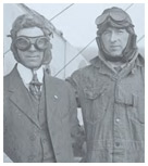 Black and white photo of twom men in aviation gear standing against a bi-plane.