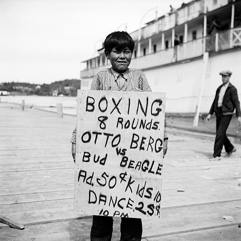 Black and white photo of a boy in a collared shirt holding a sign that reads "Boxing 8 rounds Otto Berg vs Bud Beagle Ad. 50 cents Kids 10 Dance 25 cents 10PM."