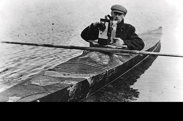 This image is greyscale and shows a nman in a kayak with a hand-crank camera. 