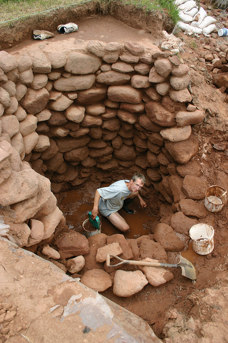 A man couches at the bottom of a rock well that is being excavated. 