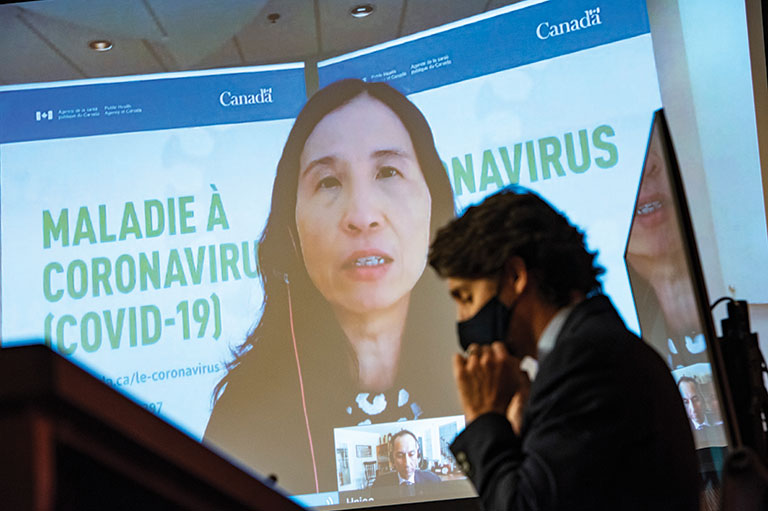A large screen displays a video of Canada's health minister Theresa Tam. Justin Trudeau, Canada's Prime Minister, sits at a table beside the screen wearing a mask.