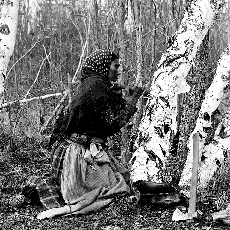 Black and white photo of a woman in a shawl and headscarf kneeling beside a tree and carving into it.