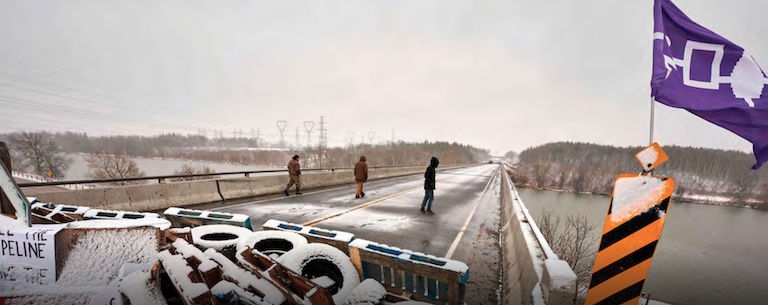 A highway road is blockaded in winter with tires and wooden pallets. Three people walk down the road and a flag with purple and white markings waves in the wind.
